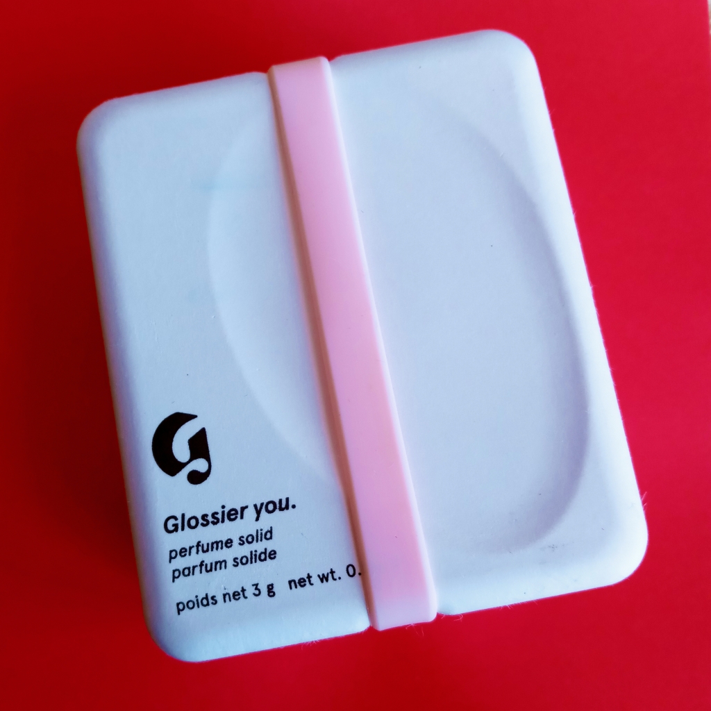 The white Glossier You solid perfume box from 2022, closed. The brand's logo and the box's contents are stamped in black ink. The pink rubber band is wrapped around the box.