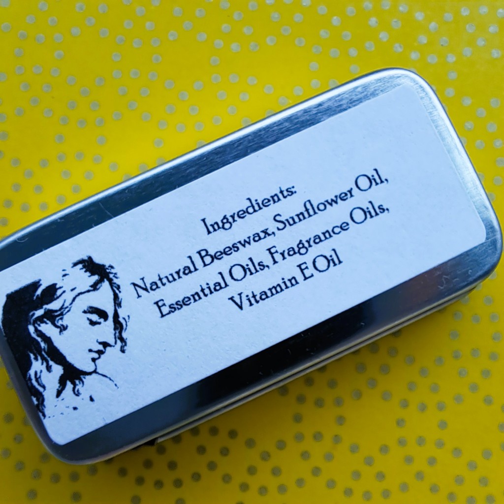 The bottom of the perfume tin, adorned wtih a classical image of a woman and the product's ingredients--beeswax, sunflower oil, essential oil, fragrance oil, vitamin E oil