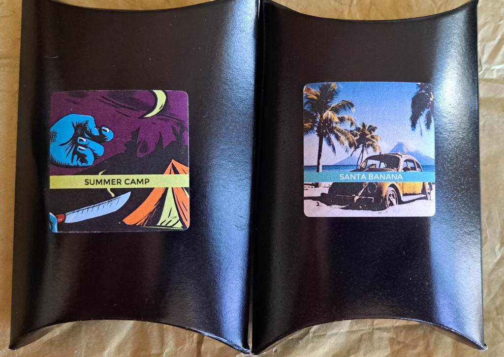 The Summper Camp box, featuring an orange tent in the moonlight, under a yellow moon, with a shadowy hand wielding a knife as someone approaches the tent, and the Santa Banana perfume box featuring a rusted-out Volkswagen Beetle with flat tires on the beach, with the ocean and palm trees in the background.