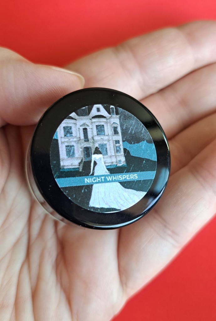 The Night Whispers perfume jar, with a black lid and a label featuring a woman in a long white dress walking through the rain towards a gloomy mansion on a hill