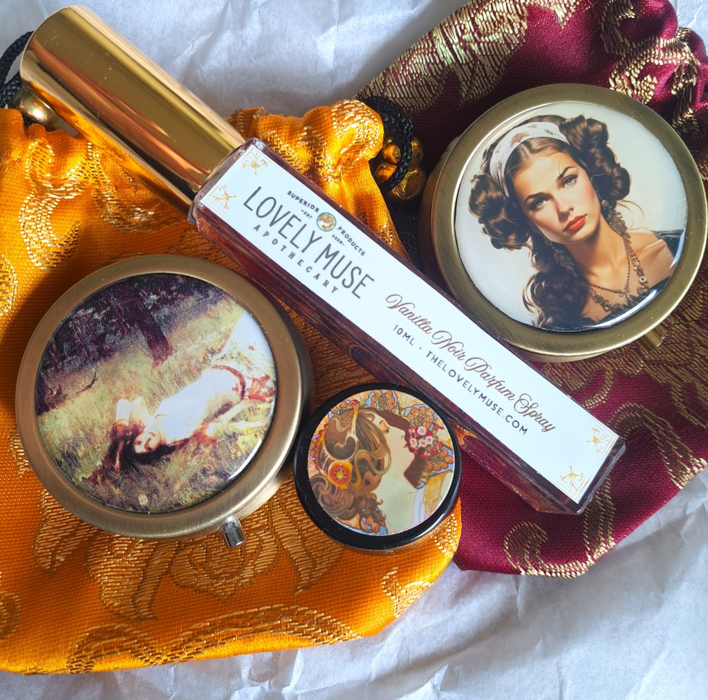 Two solid perfume compacts from The Lovely Muse Apothecary, atop their satin pouches, with a spray perfume from the brand between them, and a perfume sample from the brand resting alongside