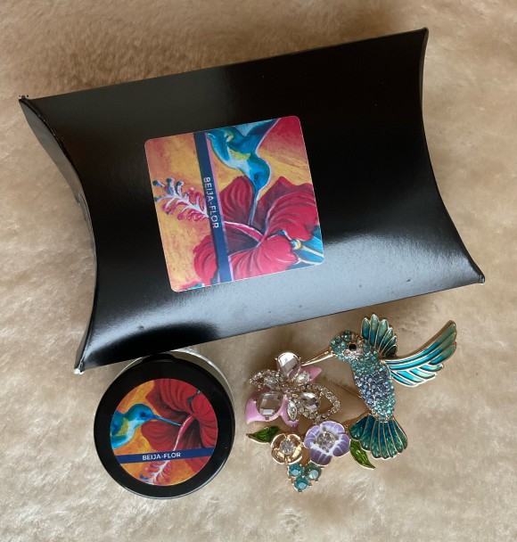 The Beija-Flor solid perfume, its pillow box, and a brooch that resembles a blue hummingbird sipping nectar from pink and purple flowers