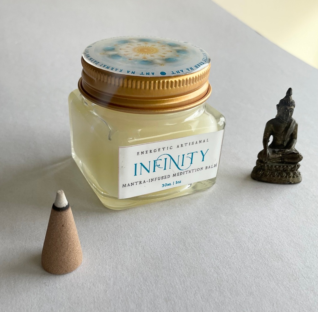 The new Infinity Meditation Balm, next to a tiny Buddha statue and a cone of burning incense with a gentle waft of smoke